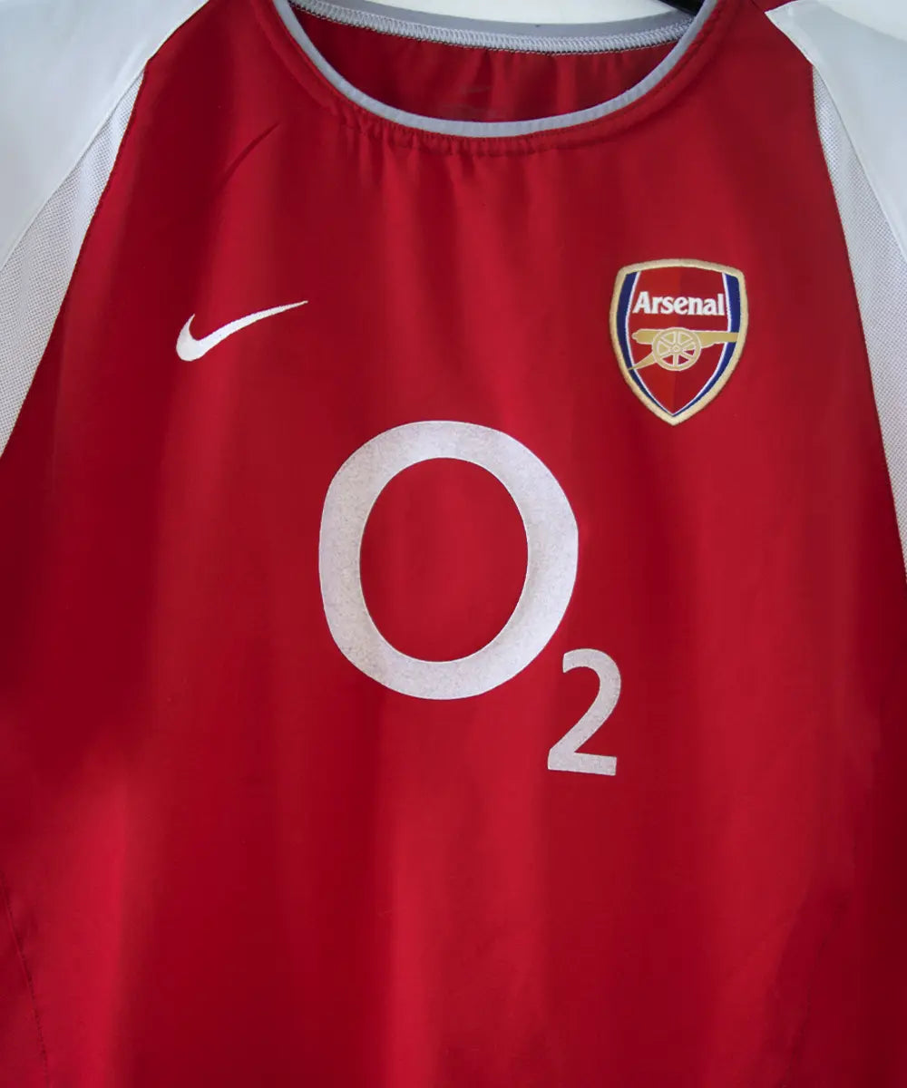 maillot authentique arsenal 2002-2004. Le maillot est de couleur rouge et blanche. On peut retrouver le sponsor O2 en feutrine et l'équipementier nike. Le maillot est floqué du numéro 14 Thierry Henry. Sur cette photo on peut voir le devant du maillot de près