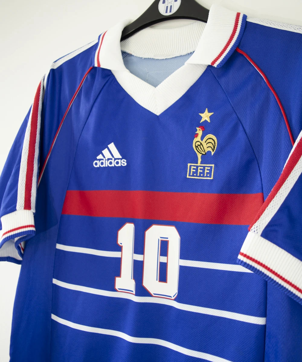 Maillot vintage domicile bleu, blanc et rouge de l'équipe de france 1998. On peut retrouver l'équipementier adidas. Le maillot est floqué du numéro 10 Zinedine Zidane avec le flocage à l'avant et à l'arrière du maillot. Il s'agit d'un maillot authentique d'époque