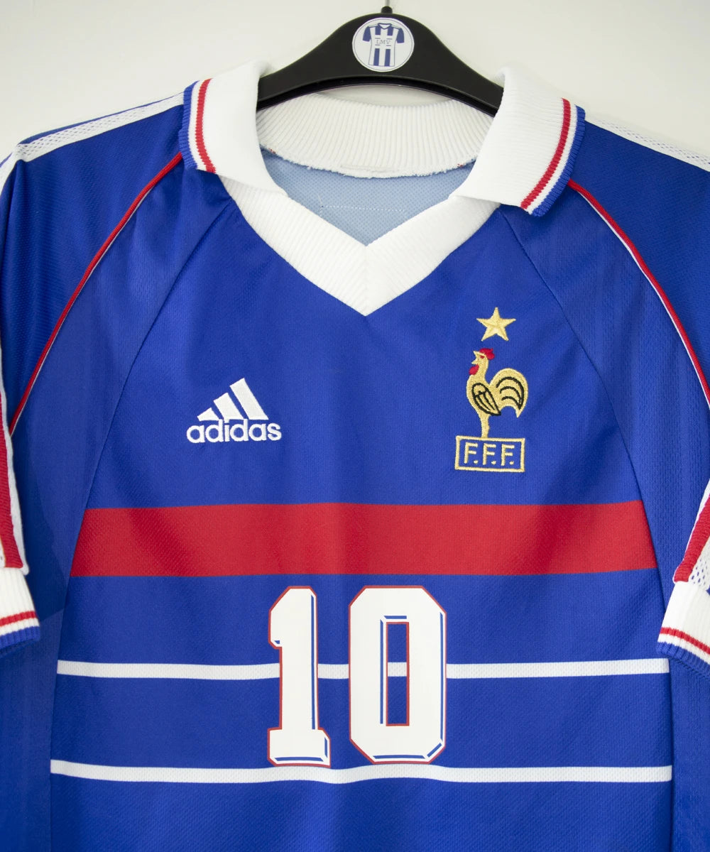 Maillot vintage domicile bleu, blanc et rouge de l'équipe de france 1998. On peut retrouver l'équipementier adidas. Le maillot est floqué du numéro 10 Zinedine Zidane avec le flocage à l'avant et à l'arrière du maillot. Il s'agit d'un maillot authentique d'époque