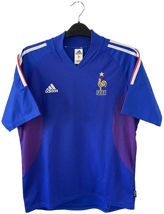 Maillot de foot vintage domicile de l'Equipe de France 2002. Le maillot est de couleur bleu, blanc et rouge. On peut retrouver l'équipementier adidas et le flocage de Zinedine Zidane. Il s'agit d'un maillot authentique. Le maillot possède l'étiquette d'authenticité comportant les numéros 139531.