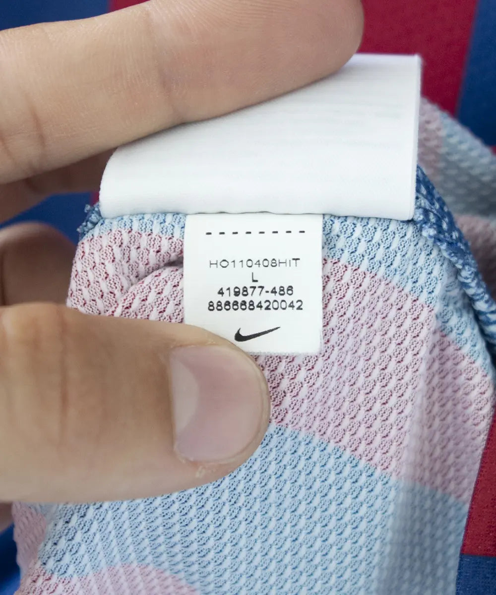 Maillot domicile bleu et rouge du fc barcelone de la saison 2011-2012. On peut retrouver le sponsor qatar foundation, le sponsor unicef et l'équipementier nike. Le maillot est floqué du numéro 10 Leo Messi. Sur cette photo on peut voir l"étiquette d'authenticité comportant les numéros 419877-486
