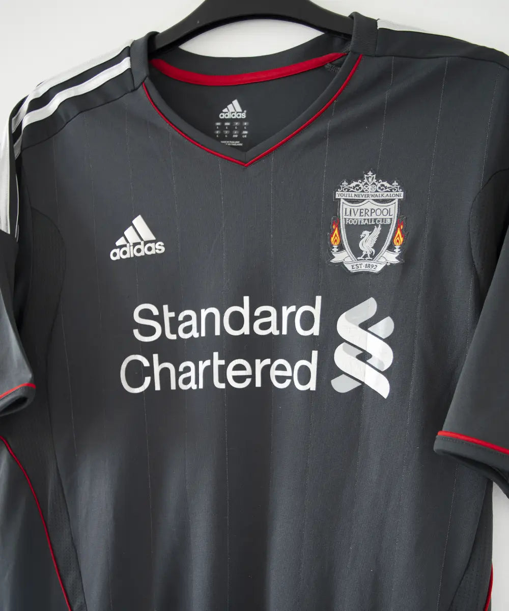 Maillot extérieur de liverpool de la saison 2011-2012. Le maillot est de couleur noir, gris et rouge. On peut retrouver l'équipementier adidas et le sponsor standard chartered.