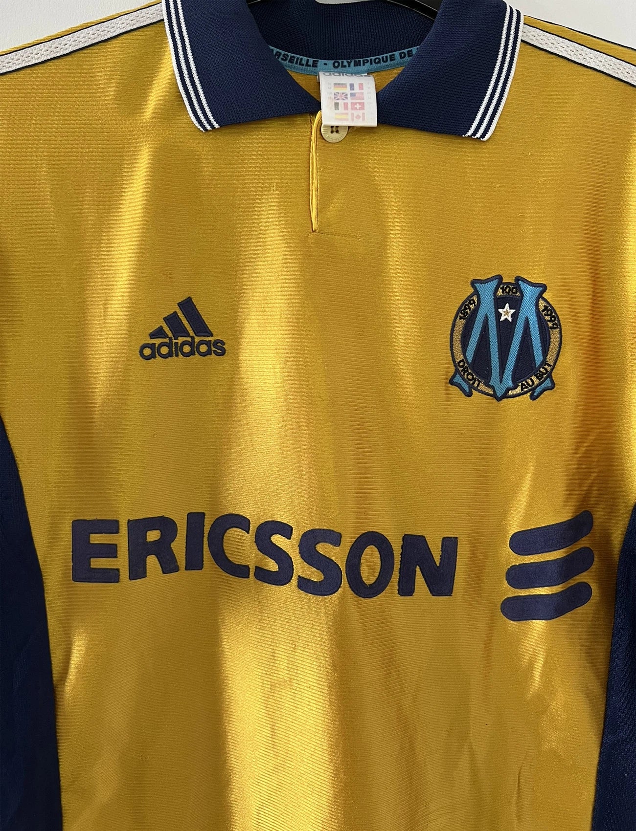 Maillot de foot vintage third de l'OM de la saison 1998-1999. Il s'agit du maillot édité pour le centenaire du club. Le maillot est de couleur dorée et bleu. On peut retrouver l'équipementier adidas et le sponsor ericsson. Il s'agit d'un maillot authentique d'époque.
