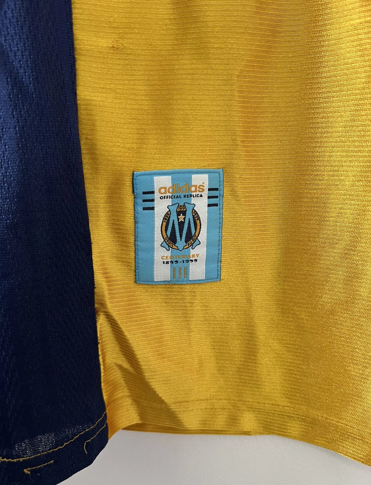 Maillot de foot vintage third de l'OM de la saison 1998-1999. Il s'agit du maillot édité pour le centenaire du club. Le maillot est de couleur dorée et bleu. On peut retrouver l'équipementier adidas et le sponsor ericsson. Il s'agit d'un maillot authentique d'époque.