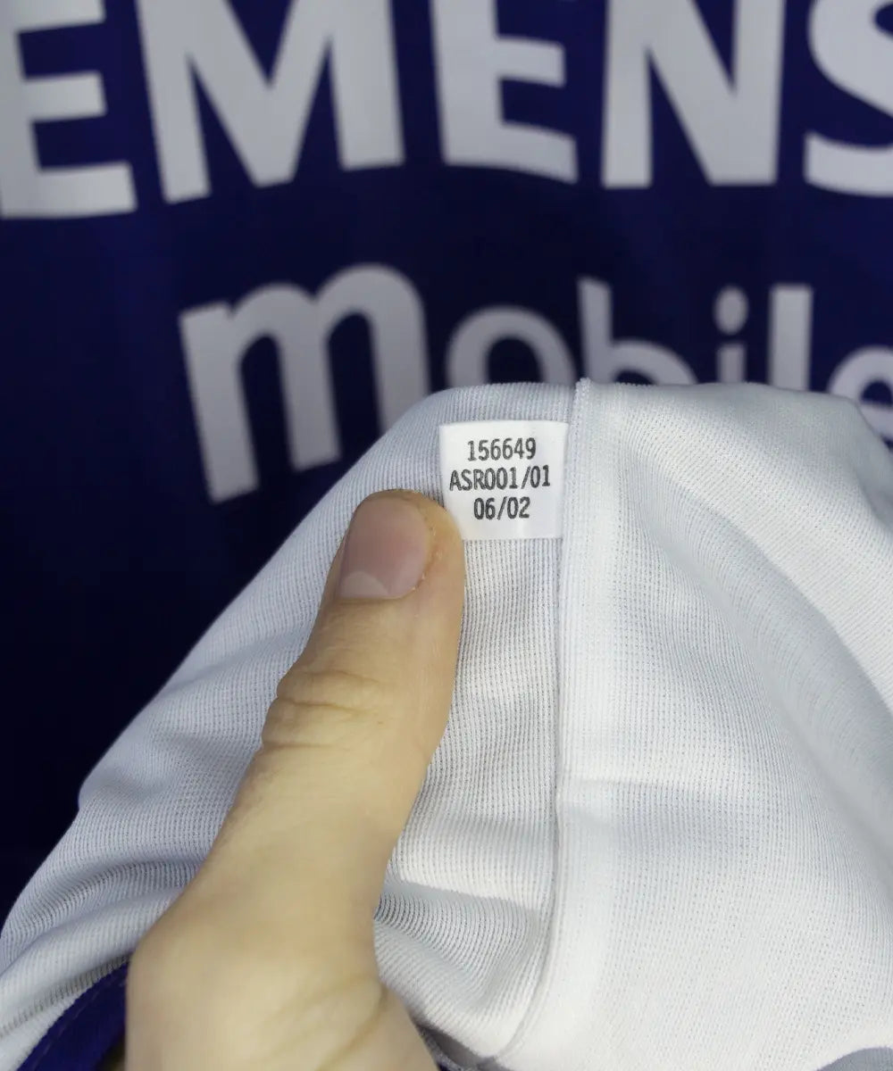 Maillot vintage violet et blanc third du real madrid de la saison 2002-2003. On peut retrouver l'équipementier adidas et le sponsor siemens mobile. Il s'agit du maillot du centenaire. Le maillot est floqué du numéro 5 Zinedine Zidane. Sur cette photo on peut voir l'étiquette d'authenticité comportant les numéros 156649