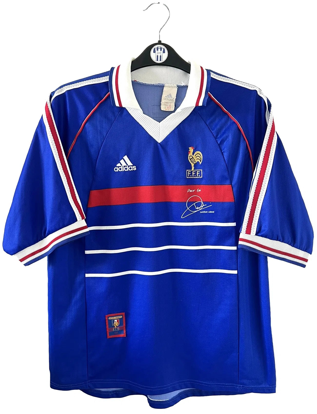 Maillot de foot vintage de l'équipe de france bleu blanc et rouge. Il s'agit du maillot domicile de la coupe du monde 1998. On peut retrouver l'équipementier adidas et le coq sans l'étoile. Le maillot possède la signature pour toi de Zinedine Zidane