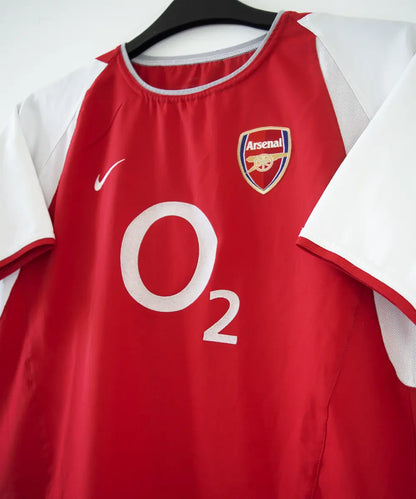 maillot authentique arsenal 2002-2004. Le maillot est de couleur rouge et blanche. On peut retrouver le sponsor O2 en feutrine et l'équipementier nike. Le maillot est floqué du numéro 14 Thierry Henry. Sur cette photo on peut voir le devant du maillot de côté