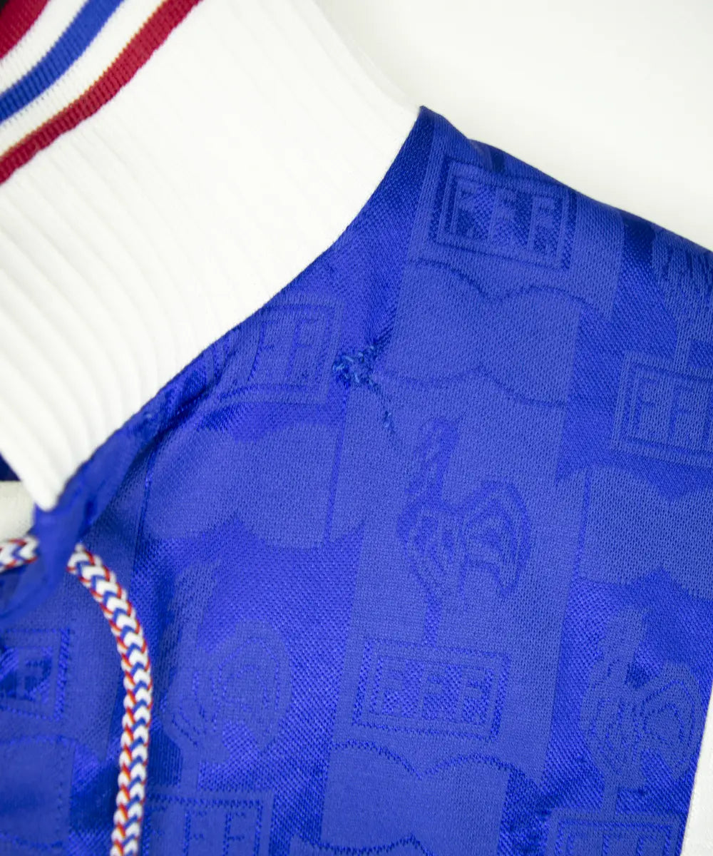 Maillot domicile de l'équipe de france 1996 de couleur bleu blanc et rouge. On peut retrouver l'équipementier adidas et le coq sans étoile