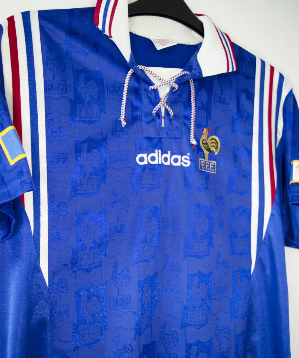 Maillot domicile de l'équipe de france 1996 de couleur bleu blanc et rouge. On peut retrouver l'équipementier adidas et le coq sans étoile