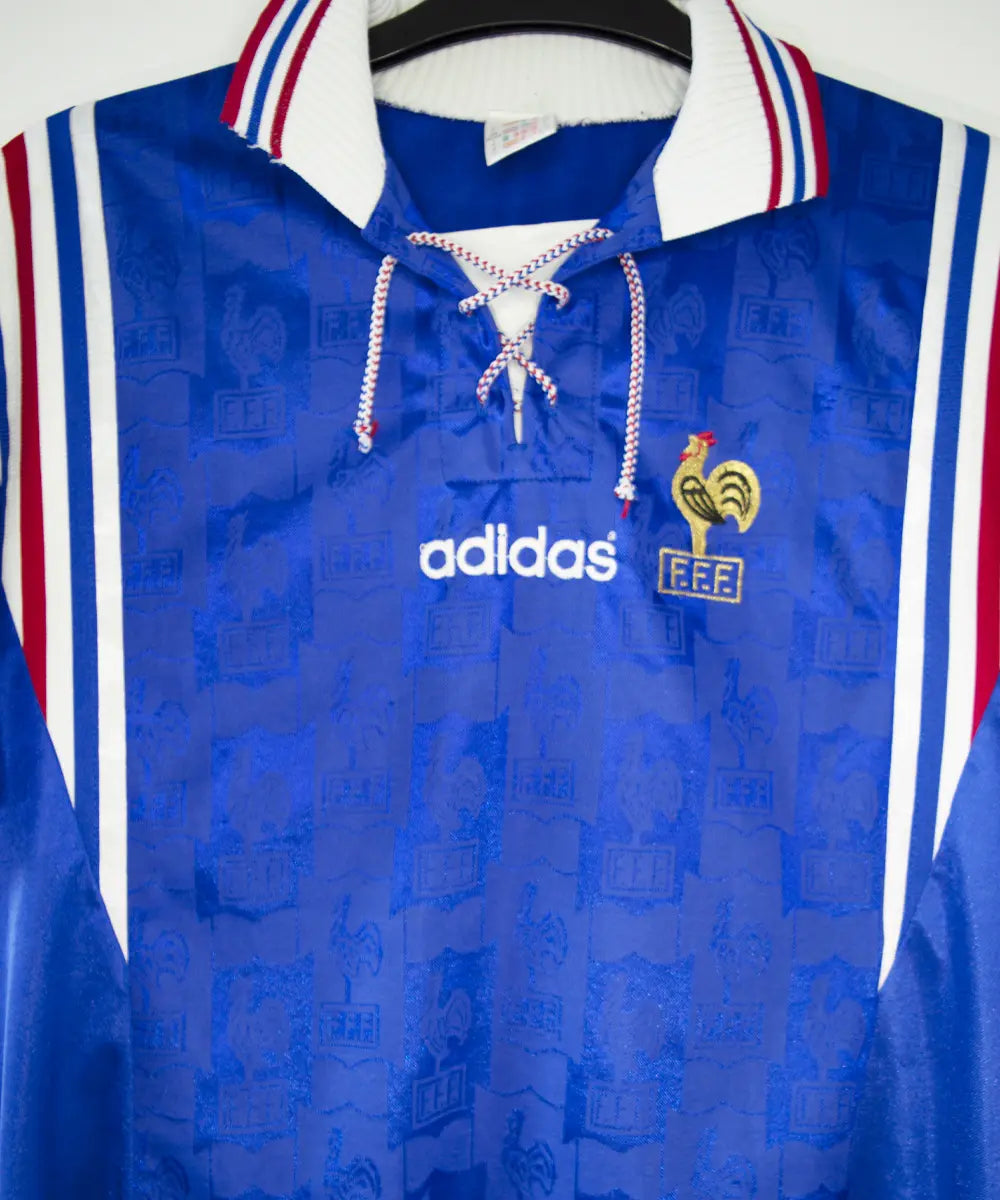 Maillot domicile de l'équipe de france 1996 de couleur bleu blanc et rouge. On peut retrouver l'équipementier adidas et le coq sans étoile