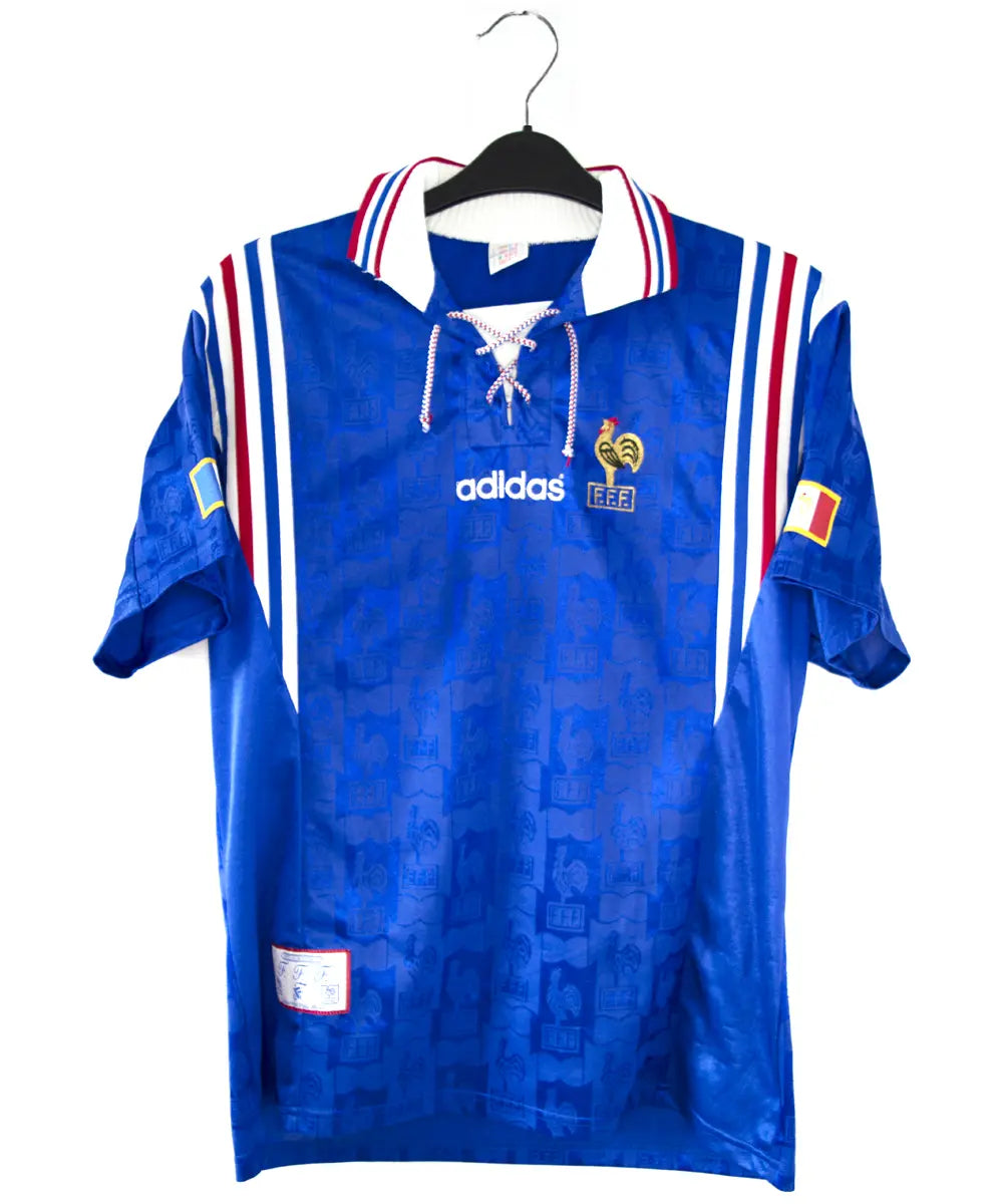 Maillot domicile de l'équipe de france 1996 de couleur bleu blanc et rouge. On peut retrouver l'équipementier adidas et le coq sans étoile