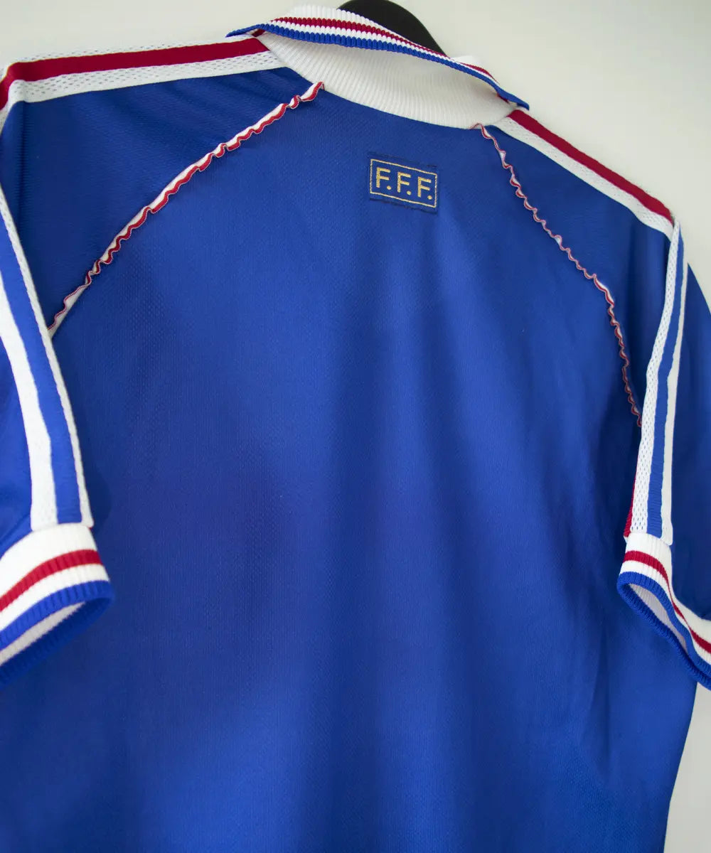 Maillot domicile de l'équipe de france 1998. Le maillot est de couleur bleu, blanc et rouge. On peut retrouver l'équipementier adidas et le coq sans étoile. Le maillot est signé Didier Deschamps