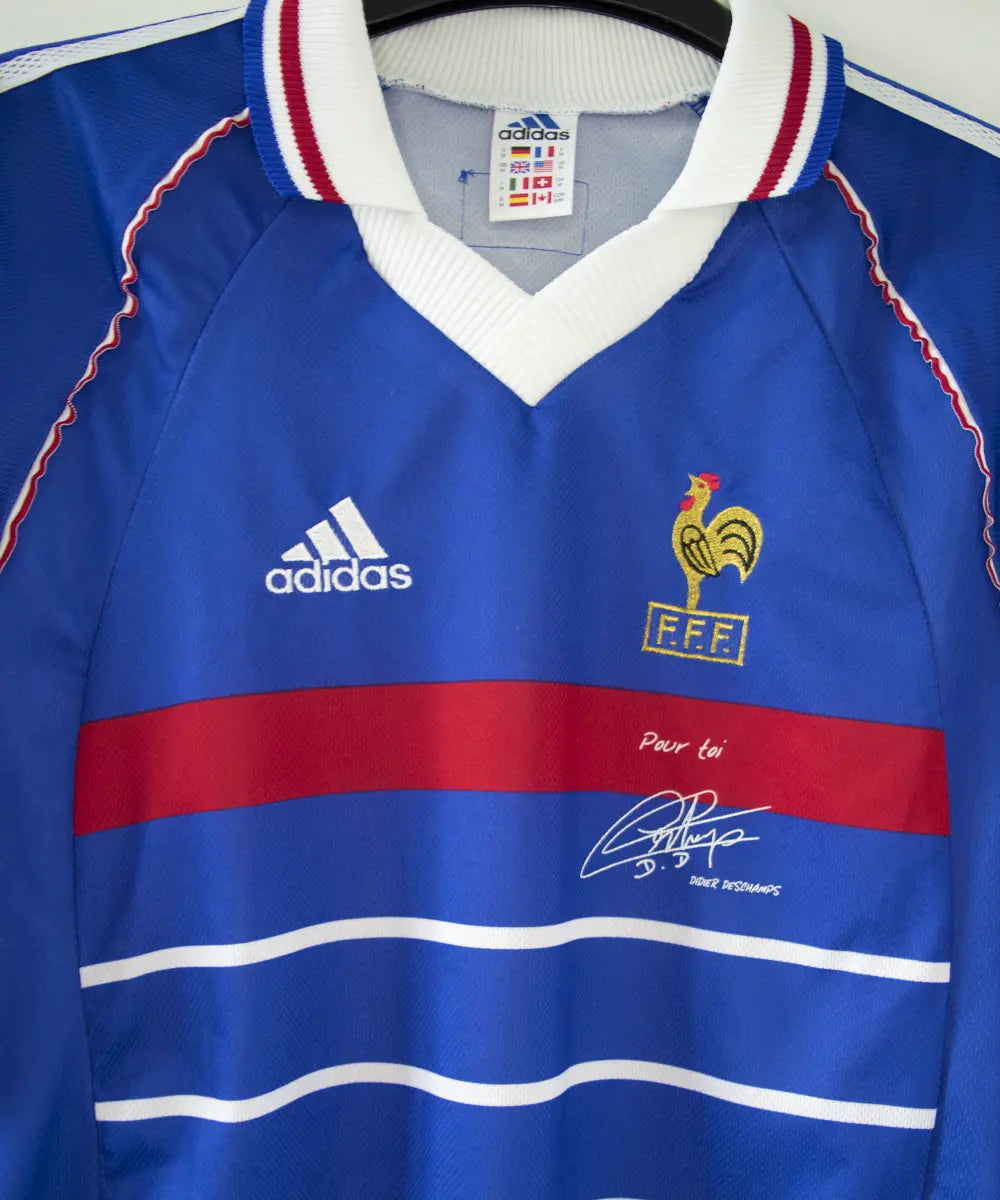 Maillot domicile de l'équipe de france 1998. Le maillot est de couleur bleu, blanc et rouge. On peut retrouver l'équipementier adidas et le coq sans étoile. Le maillot est signé Didier Deschamps