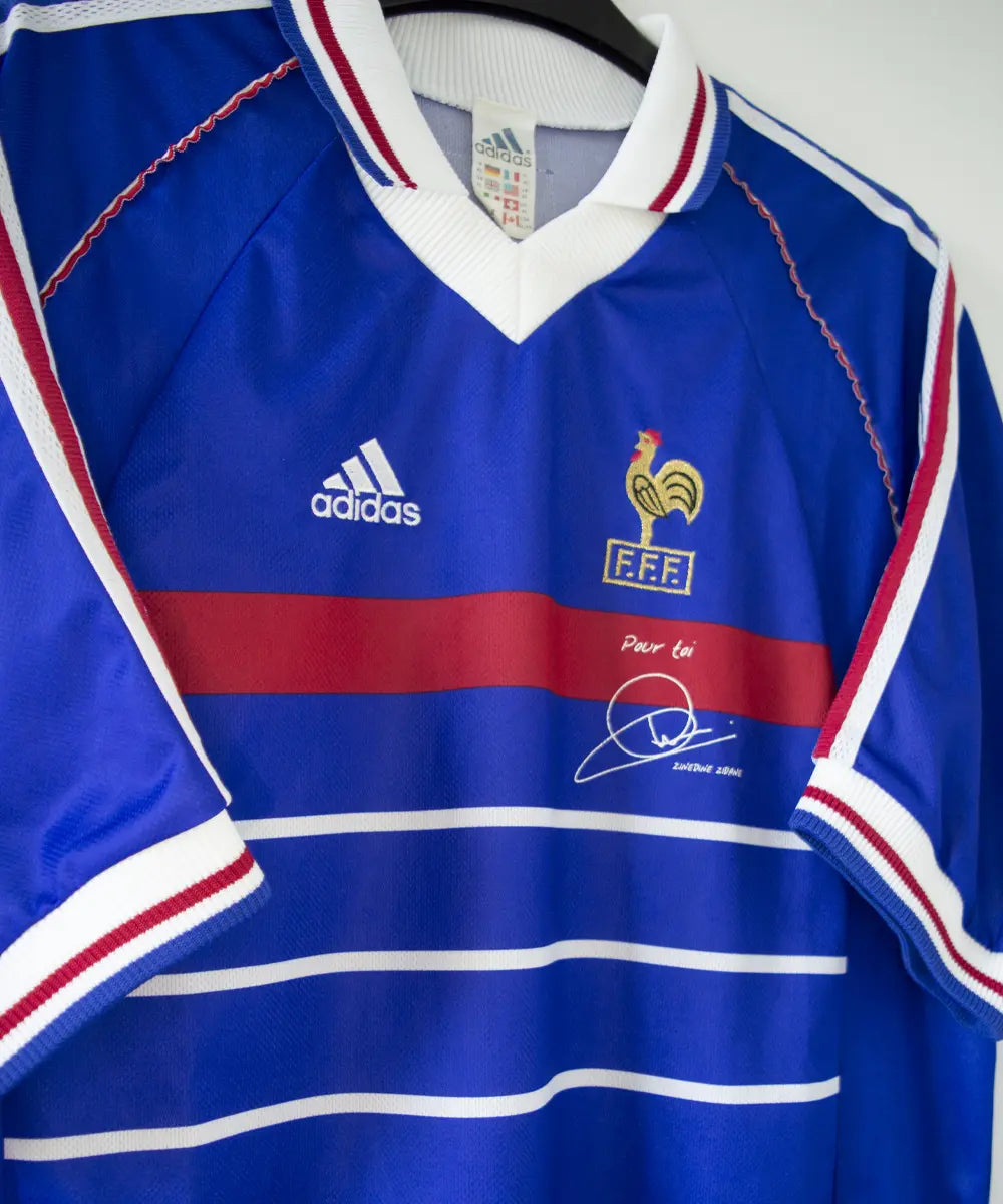 Maillot domicile équipe de france 1998 signé Zidane. Le maillot est de couleur bleu. On peut retrouver l'équipementier adidas et le coq sans étoile. Sur cette photo on peut apercevoir le maillot de côté