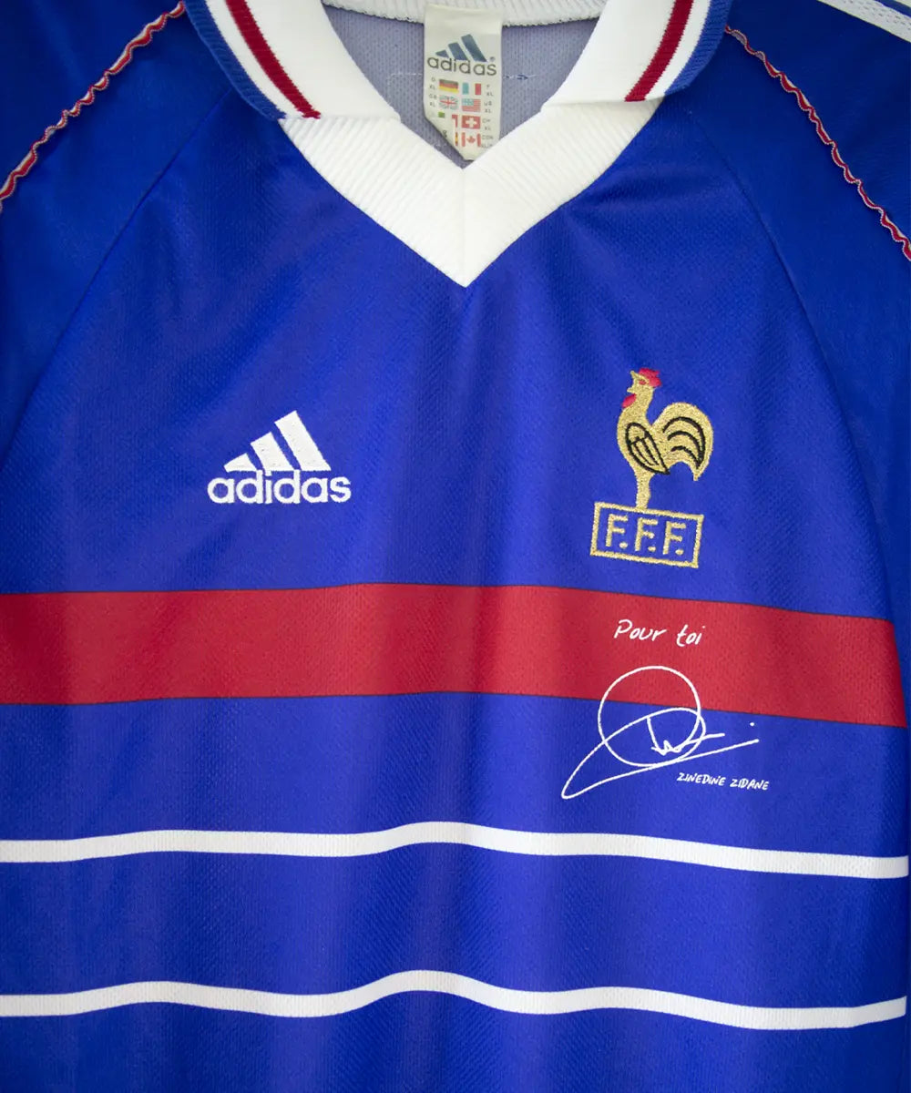 Maillot domicile équipe de france 1998 signé Zidane. Le maillot est de couleur bleu. On peut retrouver l'équipementier adidas et le coq sans étoile. Sur cette photo on peut apercevoir le maillot de face