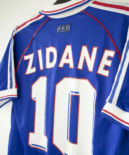 Maillot vintage domicile bleu, blanc et rouge de l'équipe de france 1998. On peut retrouver l'équipementier adidas. Le maillot est floqué du numéro 10 Zinedine Zidane avec le flocage à l'avant et à l'arrière du maillot. Il s'agit d'un maillot authentique d'époque