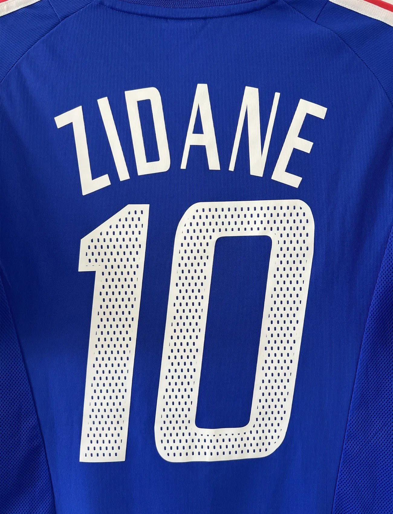 Maillot de foot vintage domicile de l'Equipe de France 2002. Le maillot est de couleur bleu, blanc et rouge. On peut retrouver l'équipementier adidas et le flocage de Zinedine Zidane. Il s'agit d'un maillot authentique. Le maillot possède l'étiquette d'authenticité comportant les numéros 139531.