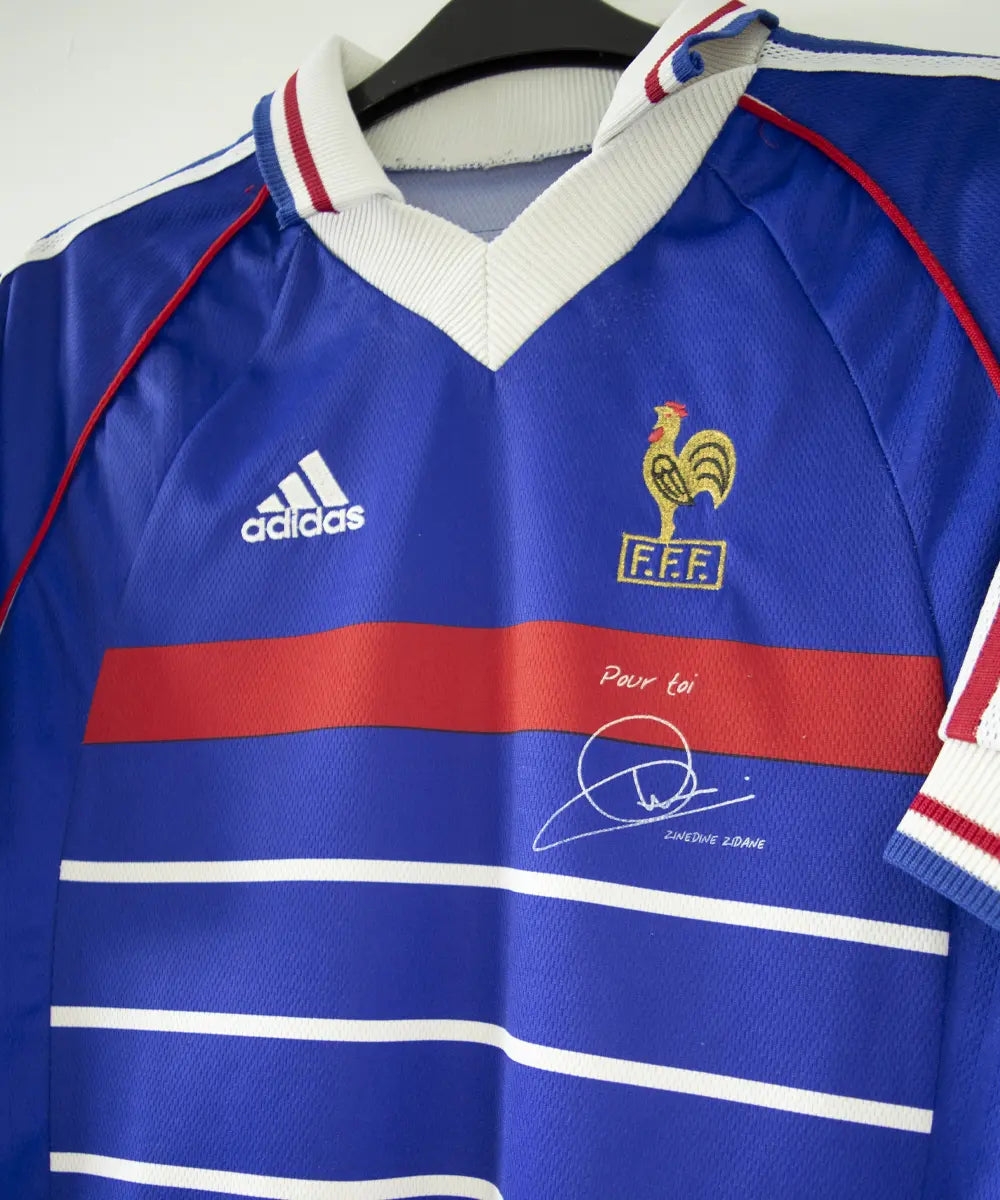 Maillot de foot de l'équipe de france 1998. Le maillot est de couleur bleu, blanc rouge. On peut retrouver l'équipementier adidas et la signature de zinedine zidane. Sur cette image on peut voir le maillot de côté