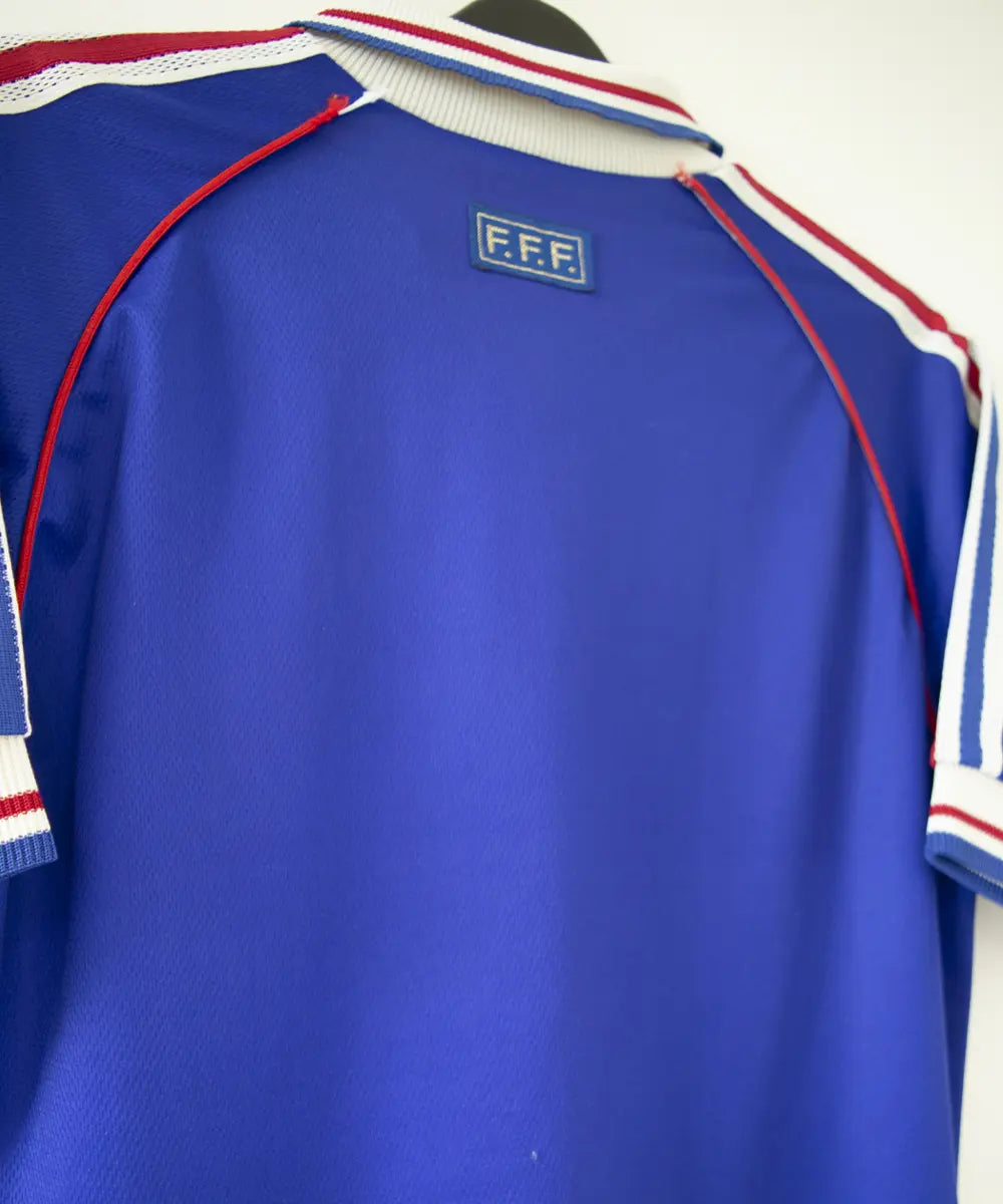 Maillot de foot de l'équipe de france 1998. Le maillot est de couleur bleu, blanc rouge. On peut retrouver l'équipementier adidas et la signature de zinedine zidane. Sur cette photo on peut voir le maillot de dos de côté