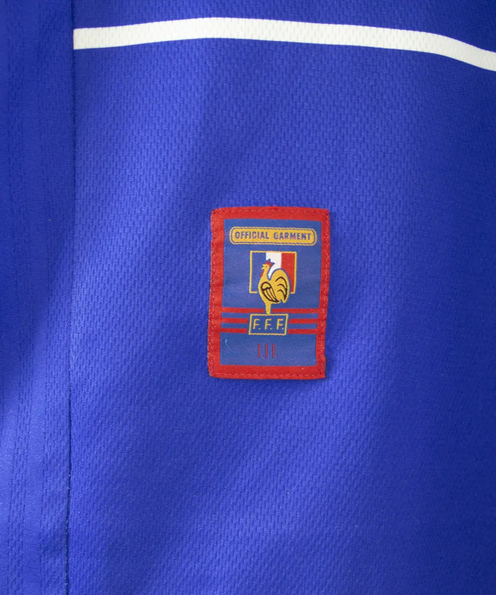 Maillot de foot de l'équipe de france 1998. Le maillot est de couleur bleu, blanc rouge. On peut retrouver l'équipementier adidas et la signature de zinedine zidane. Sur cette photo on peut voir le patch official garment