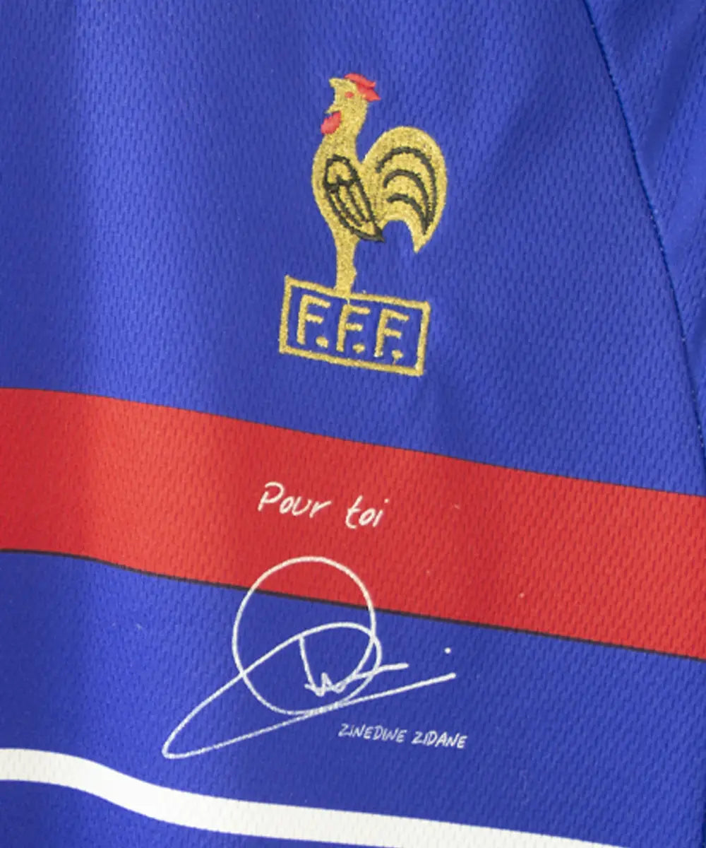 Maillot de foot de l'équipe de france 1998. Le maillot est de couleur bleu, blanc rouge. On peut retrouver l'équipementier adidas et la signature de zinedine zidane. Sur cette photo on peut voir le coq sans l'étoile et avec la signature de zidane