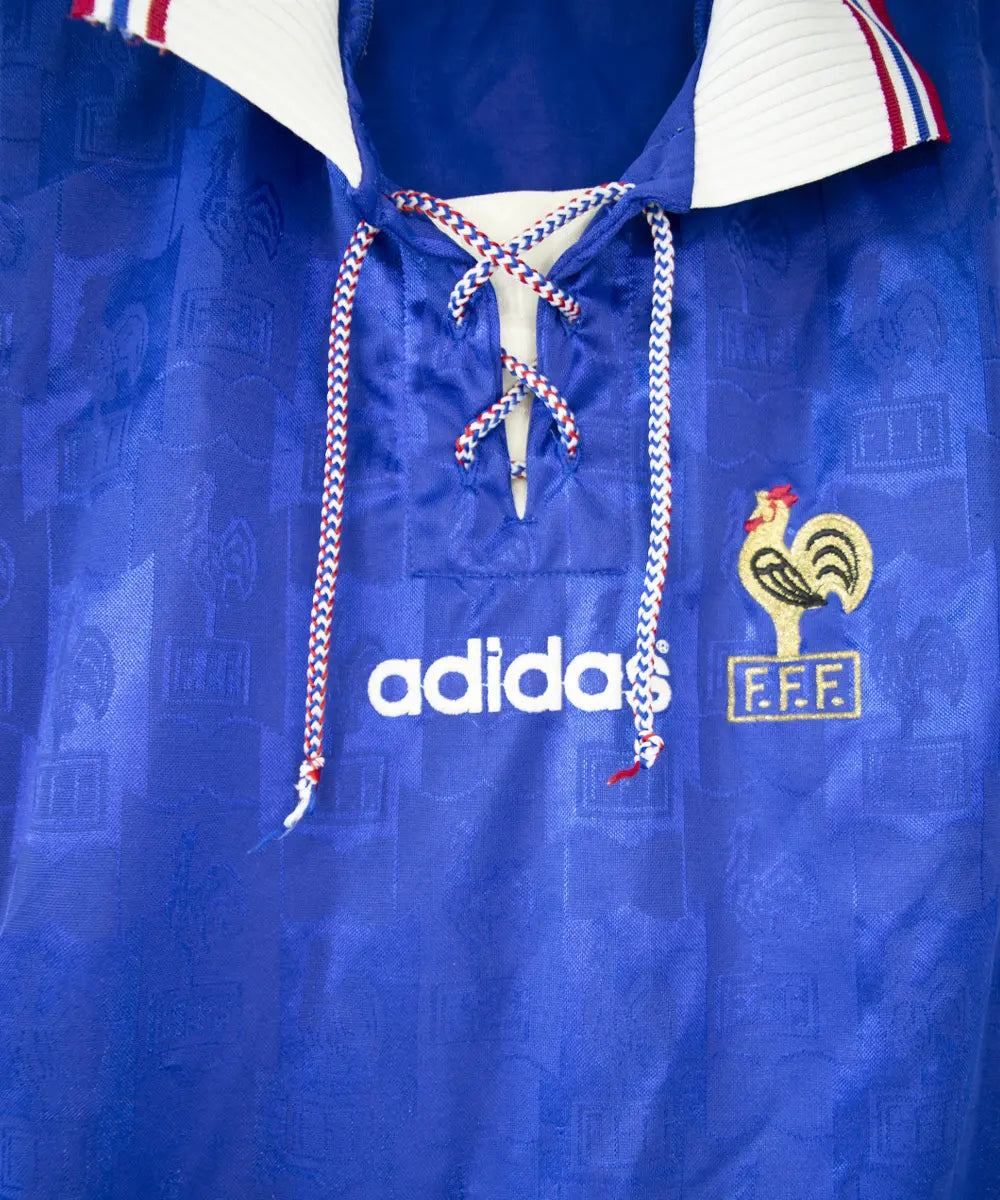 Maillot domicile bleu blanc et rouge de l'équipe de france 1996. On peut retrouver l'équipementier adidas et le coq sans l'étoile.