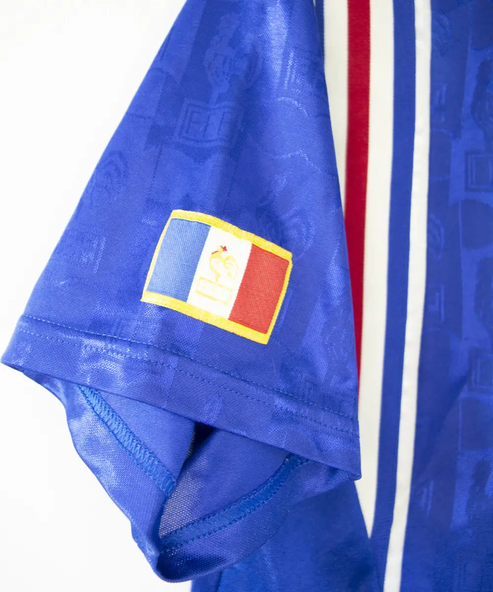 Maillot domicile bleu blanc et rouge de l'équipe de france 1996. On peut retrouver l'équipementier adidas et le coq sans l'étoile.