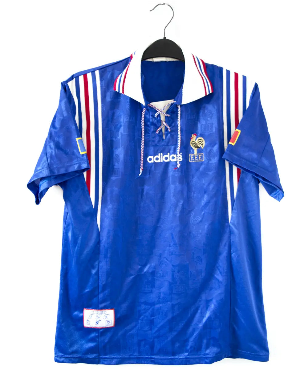 Maillot domicile bleu blanc et rouge de l'équipe de france 1996. On peut retrouver l'équipementier adidas et le coq sans l'étoile.