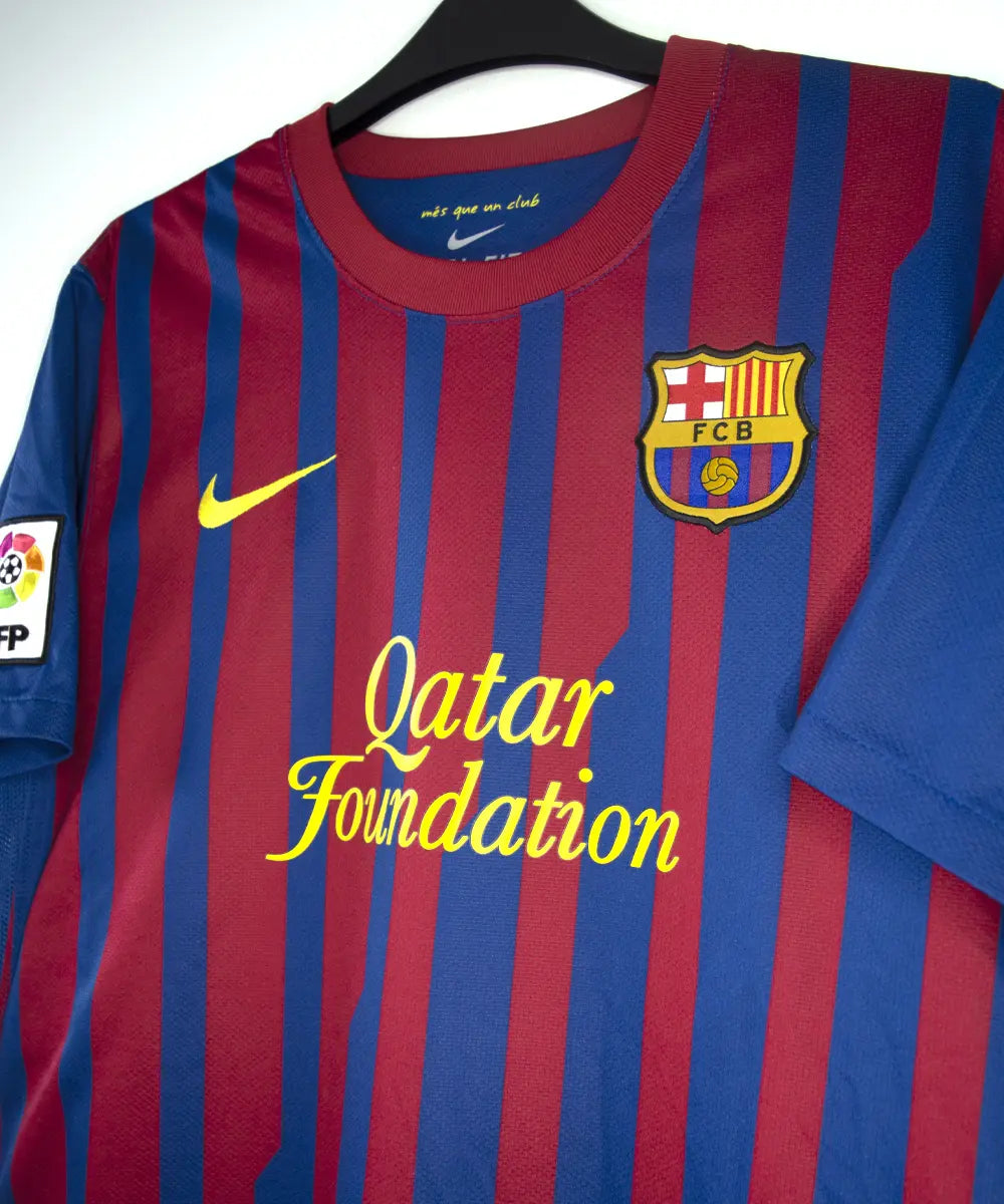 Maillot domicile bleu et rouge du fc barcelone de la saison 2011-2012. On peut retrouver le sponsor qatar foundation, le sponsor unicef et l'équipementier nike. Le maillot est floqué du numéro 10 Leo Messi. Sur cette photo on peut voir le devant du maillot de côté