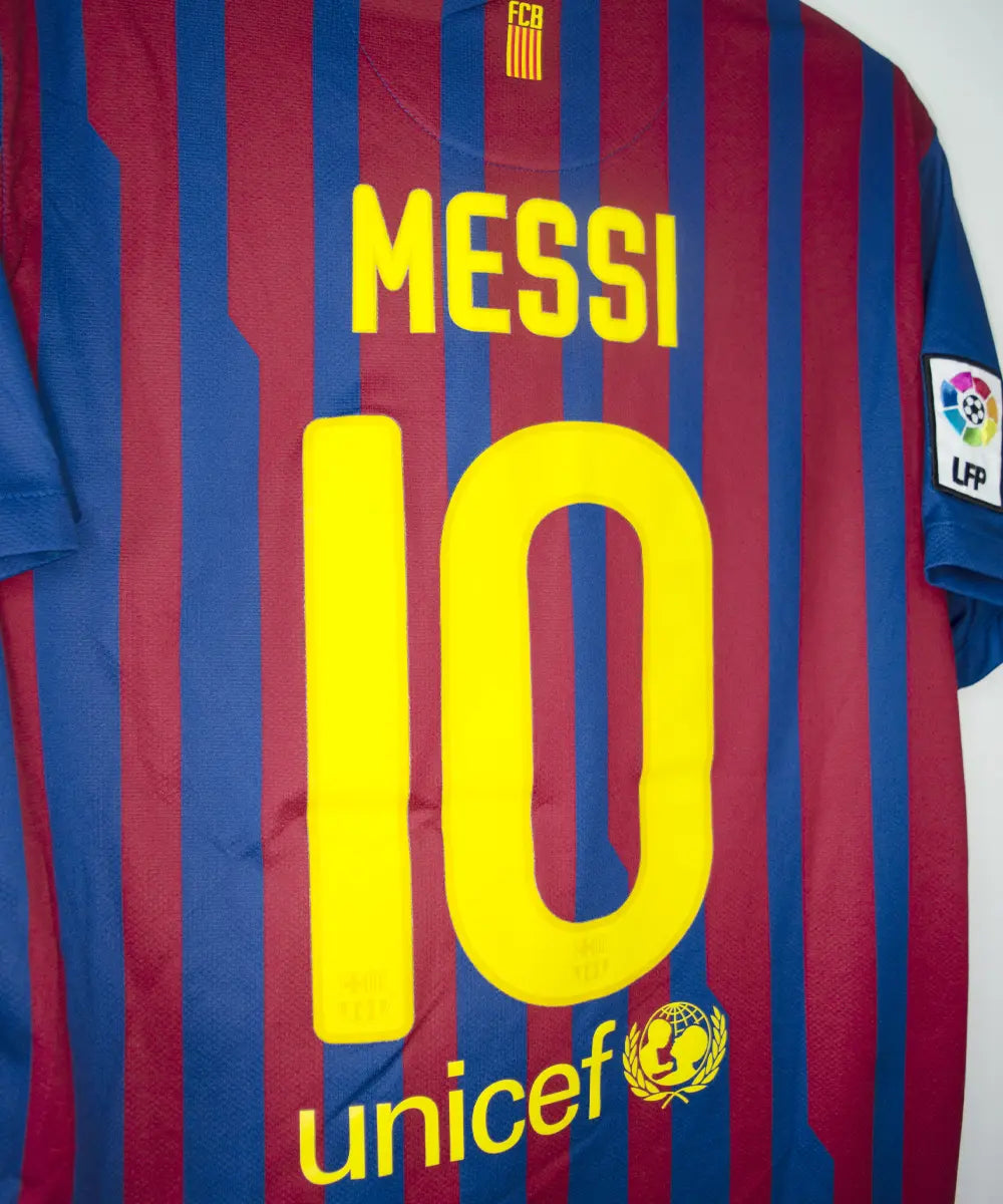 Maillot domicile bleu et rouge du fc barcelone de la saison 2011-2012. On peut retrouver le sponsor qatar foundation, le sponsor unicef et l'équipementier nike. Le maillot est floqué du numéro 10 Leo Messi. Sur cette photo on peut voir le flocage de côté
