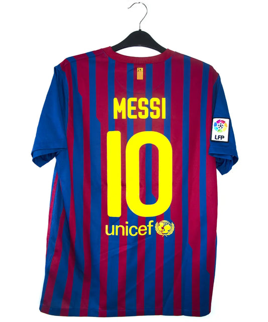 Maillot domicile bleu et rouge du fc barcelone de la saison 2011-2012. On peut retrouver le sponsor qatar foundation, le sponsor unicef et l'équipementier nike. Le maillot est floqué du numéro 10 Leo Messi. Sur cette photo on peut voir le flocage