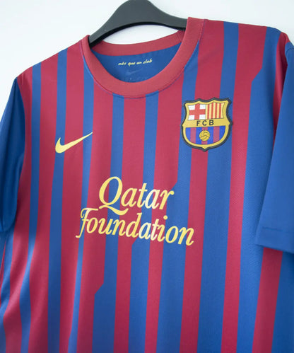 Maillot domicile bleu et rouge du fc barcelone de la saison 2011-2012. On peut retrouver le sponsor qatar foundation, le sponsor unicef et l'équipementier nike. Le maillot est floqué du numéro 10 Leo Messi. Sur cette photo on peut voir le devant du maillot de côté