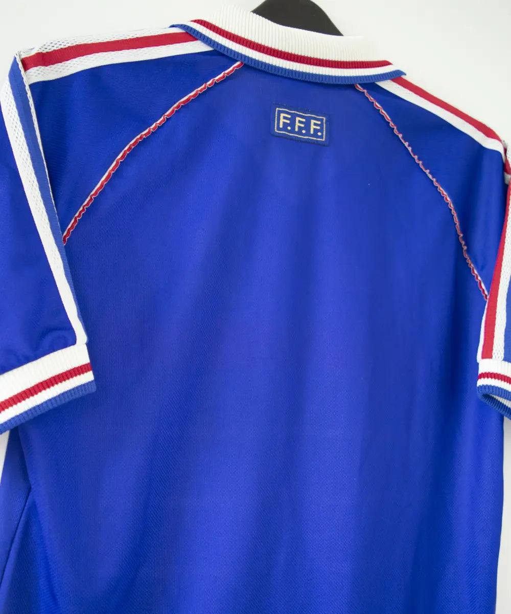 Maillot domicile bleu blanc rouge de l'équipe de france 1998. On peut retrouver l'équipementier adidas et le coq sans étoile, ainsi que le patch official garment. Sur cette photo on voit le maillot de dos de côté