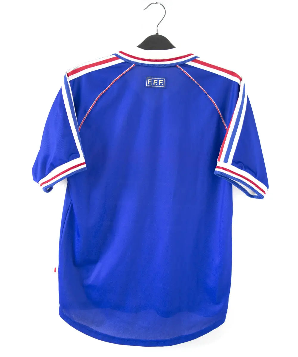 Maillot domicile bleu blanc rouge de l'équipe de france 1998. On peut retrouver l'équipementier adidas et le coq sans étoile, ainsi que le patch official garment. Sur cette photo on voit le maillot de dos
