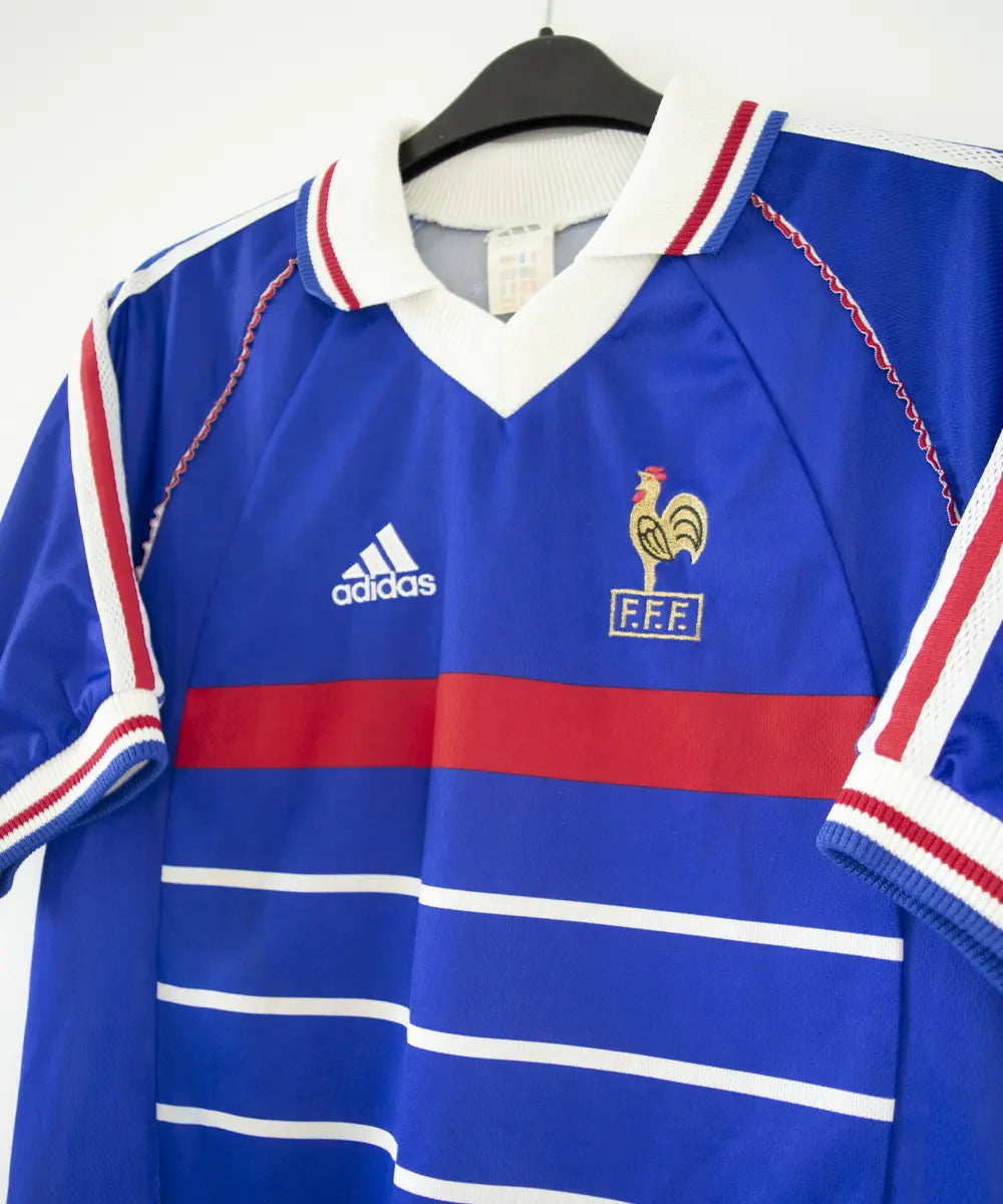 Maillot domicile bleu blanc rouge de l'équipe de france 1998. On peut retrouver l'équipementier adidas et le coq sans étoile, ainsi que le patch official garment. Sur cette photo on voit le maillot de côté