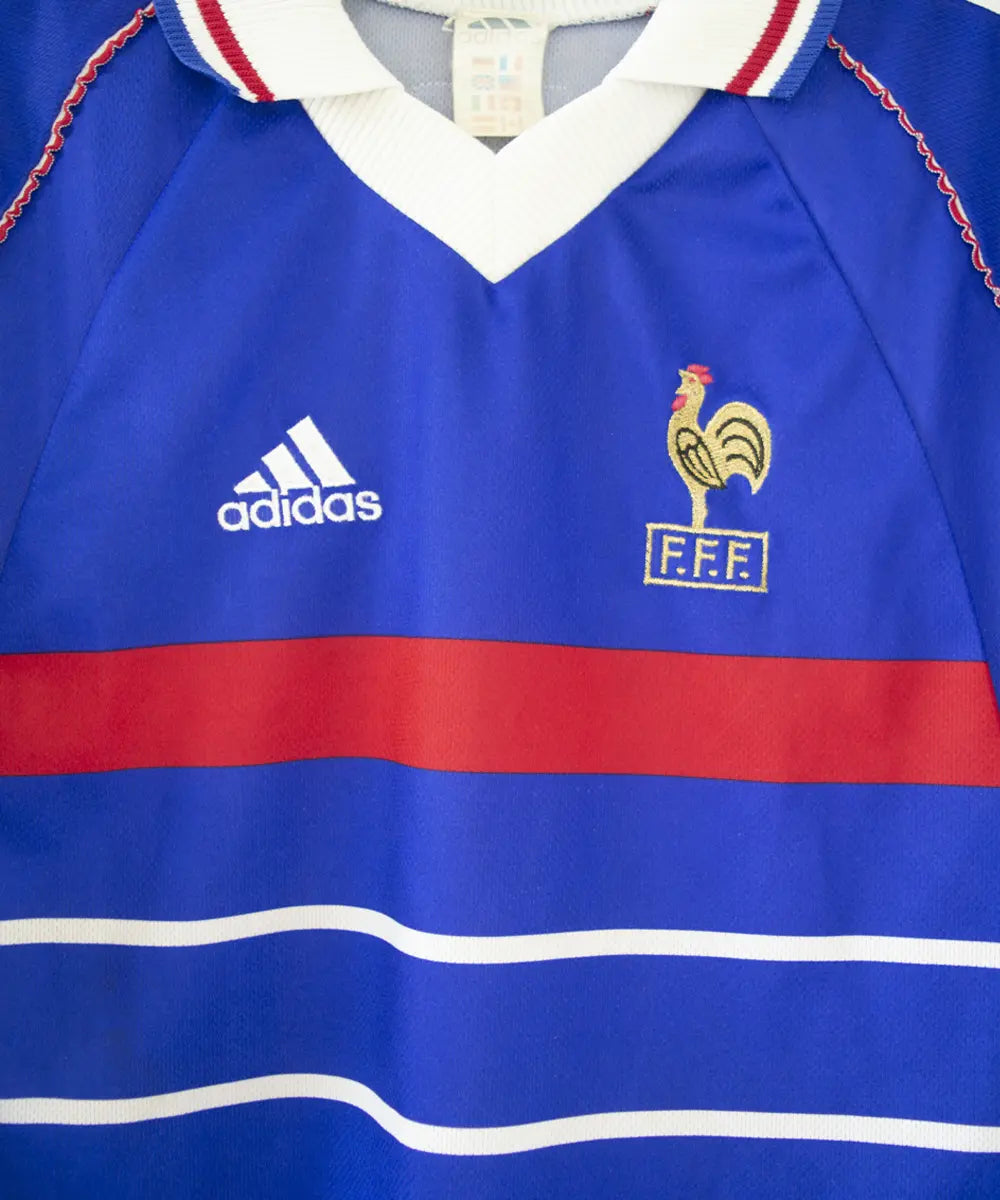 Maillot domicile bleu blanc rouge de l'équipe de france 1998. On peut retrouver l'équipementier adidas et le coq sans étoile, ainsi que le patch official garment. Sur cette photo on voit le maillot de près