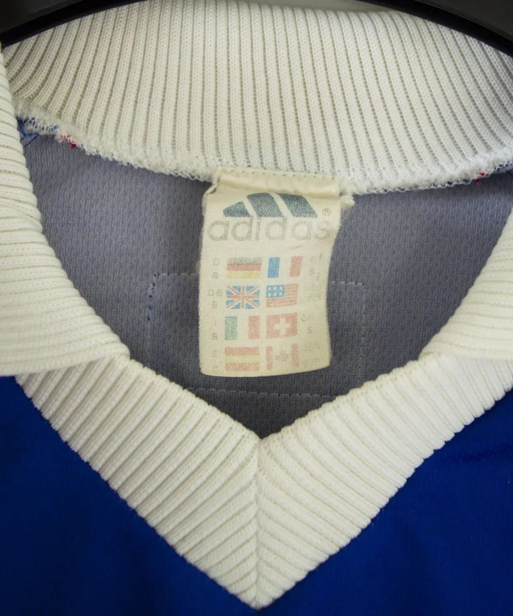 Maillot domicile bleu blanc rouge de l'équipe de france 1998. On peut retrouver l'équipementier adidas et le coq sans étoile, ainsi que le patch official garment. Sur cette photo on peut voir l'étiquette d'authenticité