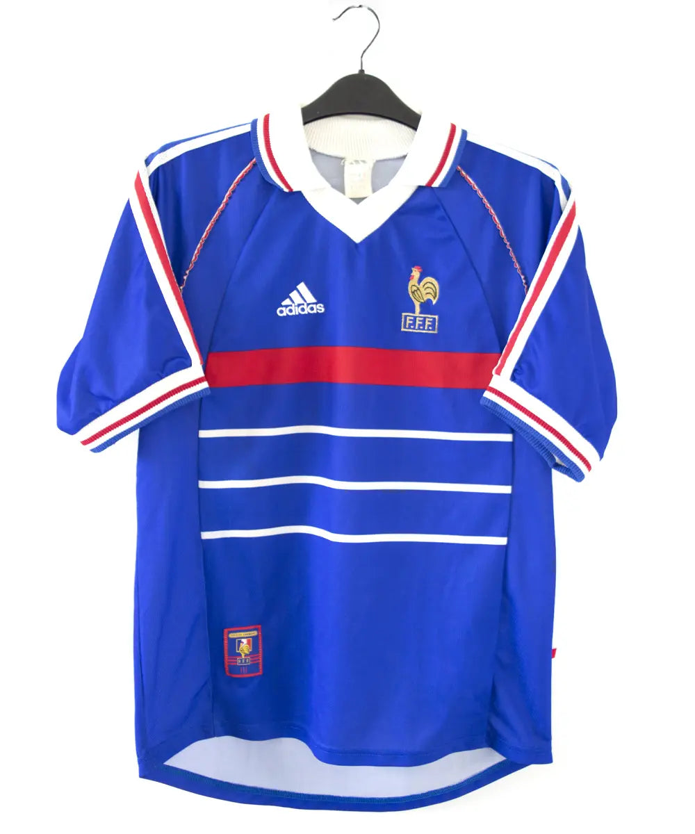 Maillot domicile bleu blanc rouge de l'équipe de france 1998. On peut retrouver l'équipementier adidas et le coq sans étoile, ainsi que le patch official garment