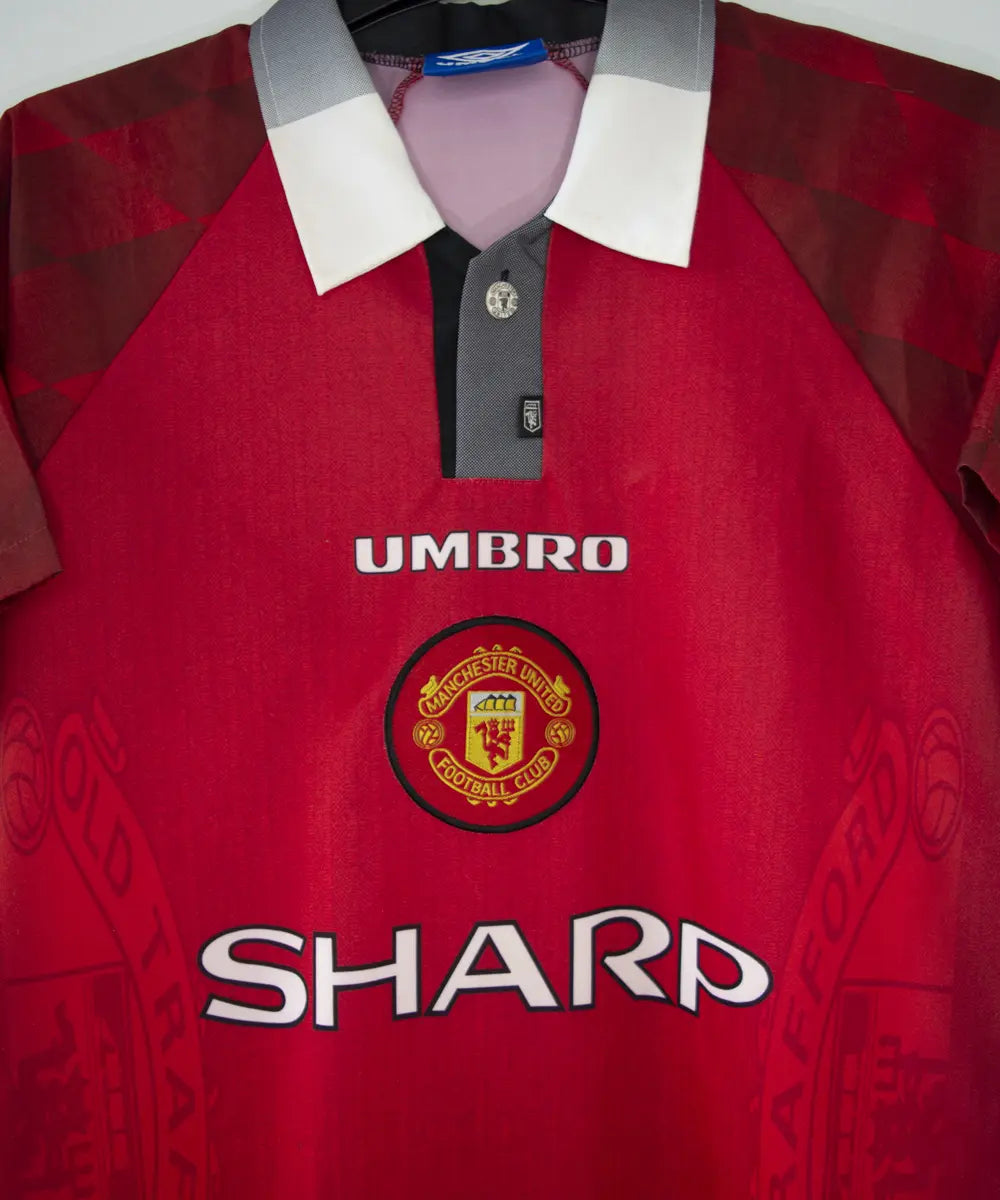 Maillot de foot vintage domicile rouge de manchester united de la saison 1996-1998. On peut retrouver l'équipementier umbro et le sponsor sharp. Le maillot est floqué du numéro 7 Cantona. Il s'agit d'un maillot authentique