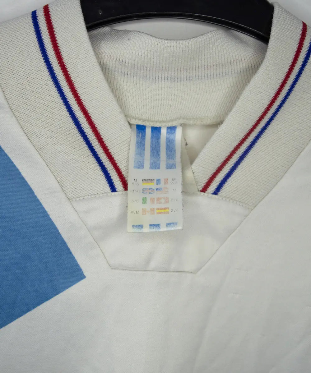 Maillot domicile blanc et bleu de l'om de la saison 1991-1992. On peut retrouver l'équipementier adidas et le sponsor Panasonic. Sur cette photo on peut voir l'étiquette adidas