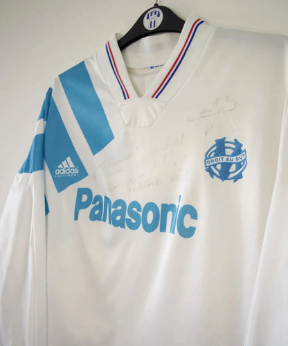 Maillot de foot vintage domicile blanc et bleu de l'OM de la saison 1991/1992. On peut retrouver l'équipementier adidas et le sponsor Panasonic. Le maillot est signé par plusieurs joueurs de l'époque dont Didier Deschamps et Jean Pierre Papin. Il s'agit d'un maillot authentique.