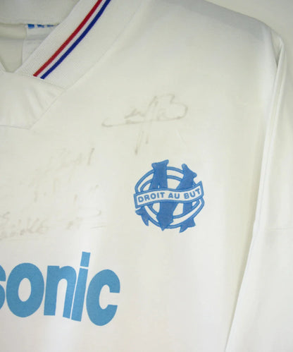 Maillot de foot vintage domicile blanc et bleu de l'OM de la saison 1991/1992. On peut retrouver l'équipementier adidas et le sponsor Panasonic. Le maillot est signé par plusieurs joueurs de l'époque dont Didier Deschamps et Jean Pierre Papin. Il s'agit d'un maillot authentique.
