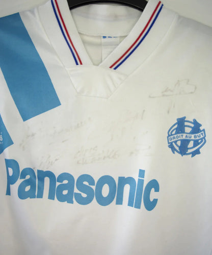 Maillot de foot vintage domicile blanc et bleu de l'OM de la saison 1991/1992. On peut retrouver l'équipementier adidas et le sponsor Panasonic. Le maillot est signé par plusieurs joueurs de l'époque dont Didier Deschamps et Jean Pierre Papin. Il s'agit d'un maillot authentique.