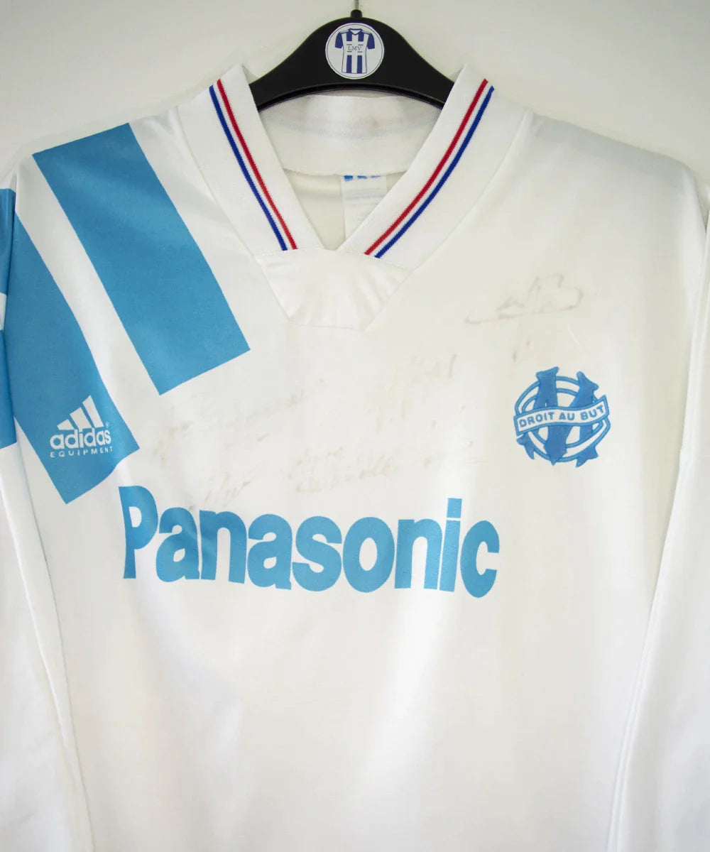 Maillot de foot vintage domicile blanc et bleu de l'OM de la saison 1991/1992. On peut retrouver l'équipementier adidas et le sponsor Panasonic. Le maillot est signé par plusieurs joueurs de l'époque dont Didier Deschamps et Jean Pierre Papin. Il s'agit d'un maillot authentique.