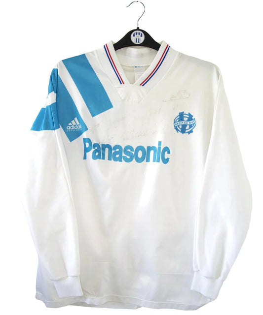Maillot de foot vintage domicile blanc et bleu de l'OM de la saison 1991/1992. On peut retrouver l'équipementier adidas et le sponsor Panasonic. Le maillot est signé par plusieurs joueurs de l'époque dont Didier Deschamps et Jean Pierre Papin. Il s'agit d'un maillot authentique.
