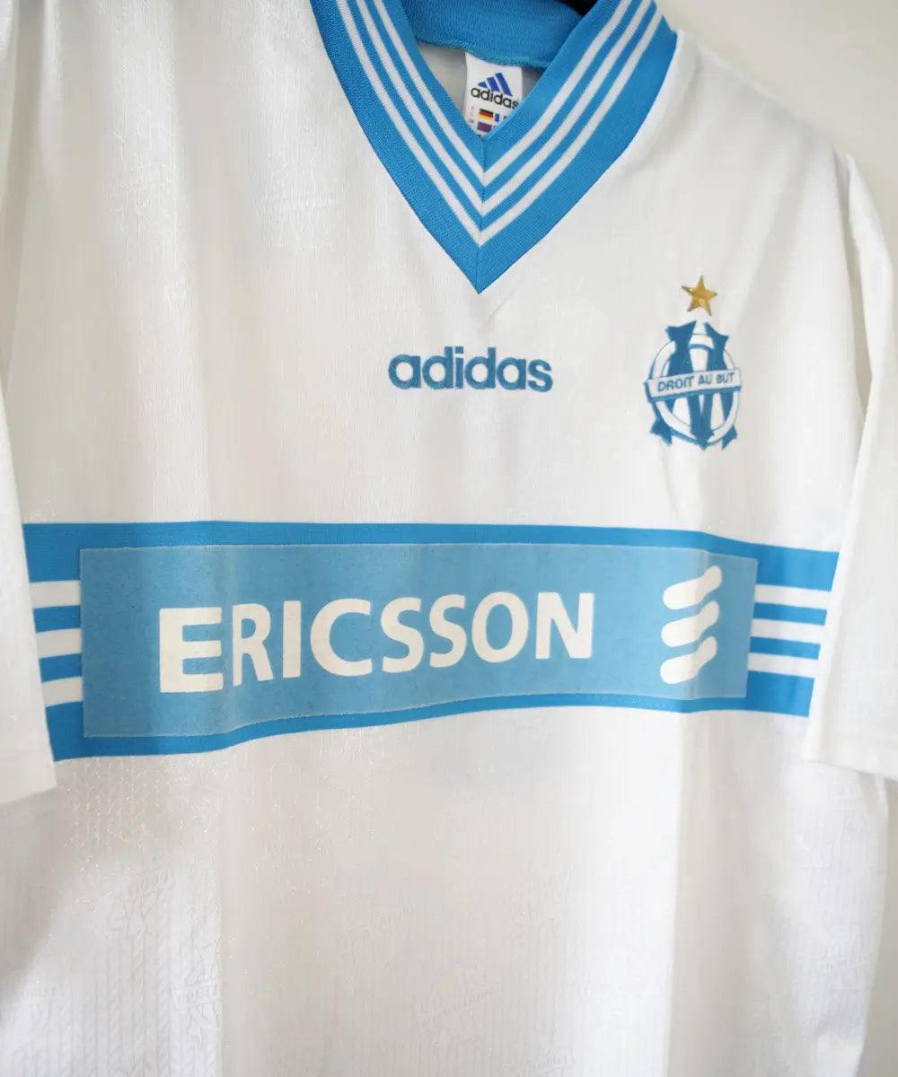 Maillot domicile blanc et bleu de l'olympique de marseille de la saison 1997-1998. Le maillot est floqué du numéro 5 Laurent Blanc. Sur cette photo on peut voir le devant du maillot avec sponsor ericsson et l'équipementier adidas. Sur cette photo on peut voir le devant du maillot de côté