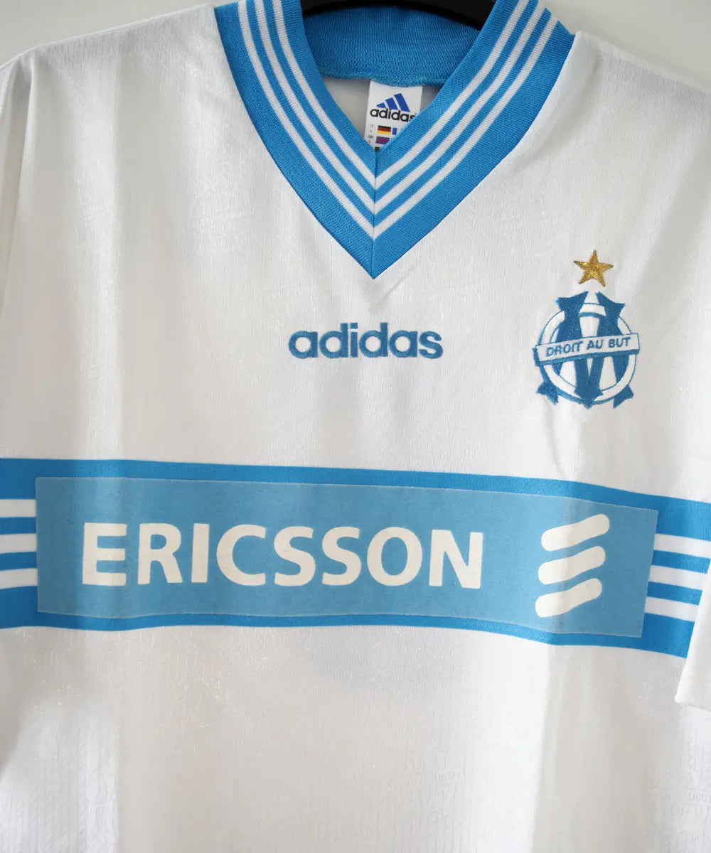 Maillot domicile blanc et bleu de l'olympique de marseille de la saison 1997-1998. Le maillot est floqué du numéro 5 Laurent Blanc. Sur cette photo on peut voir le devant du maillot avec sponsor ericsson et l'équipementier adidas. Sur cette photo on peut voir le devant du maillot de près