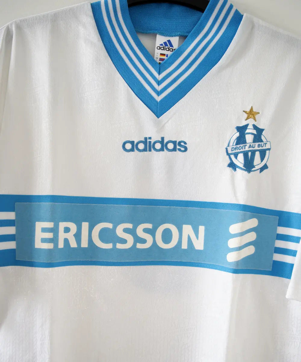 Maillot domicile blanc et bleu de l'olympique de marseille de la saison 1997-1998. Le maillot est floqué du numéro 5 Laurent Blanc. Sur cette photo on peut voir le devant du maillot avec sponsor ericsson et l'équipementier adidas. Sur cette photo on peut voir le devant du maillot de près