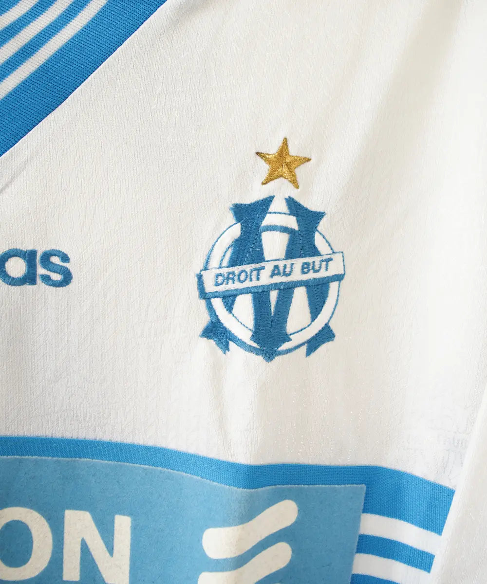 Maillot domicile blanc et bleu de l'olympique de marseille de la saison 1997-1998. Le maillot est floqué du numéro 5 Laurent Blanc. Sur cette photo on peut voir le devant du maillot avec sponsor ericsson et l'équipementier adidas. Sur cette photo on peut voir le logo brodé de l'om