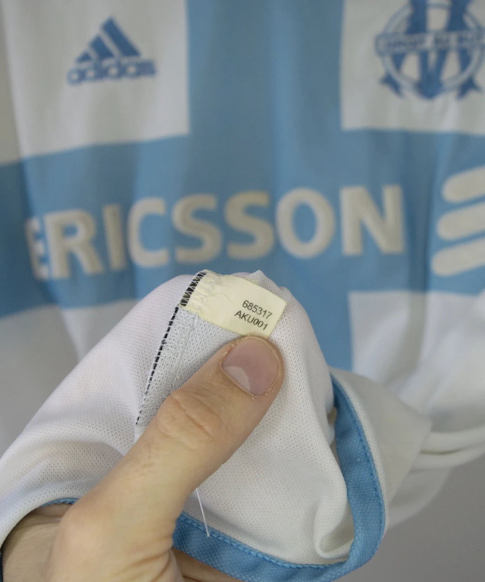 Maillot de foot vintage de l'om de la saison 2000-2001. Le maillot est de couleur blanc et bleu. On peut retrouver l'équipementier adidas et le sponsor ericsson. Ils s'agit d'un maillot authentique. On peut voir l'étiquette d'authenticité du maillot comportant les numéros 685317